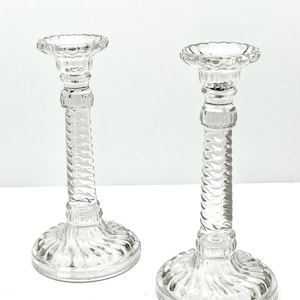 Può includere: Due candelabri in vetro trasparente con un design a spirale sullo stelo e un bordo smerlato nella parte superiore. La base è rotonda con un motivo strutturato. I candelabri sono esposti su una superficie bianca.