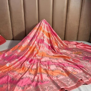 May include: A vibrant, patterned textile in shades of pink, orange, and silver. The fabric features floral and striped designs, with a detailed border. The material is draped, showcasing its flowing texture and color gradients.