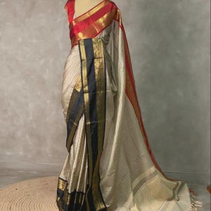 Puede incluir: Un sari color crema con finas rayas verticales, acentuado con bordes negros y dorados. El sari presenta un borde rojo y dorado a lo largo del borde y está drapeado sobre un maniquí. La blusa es roja con ribetes dorados.