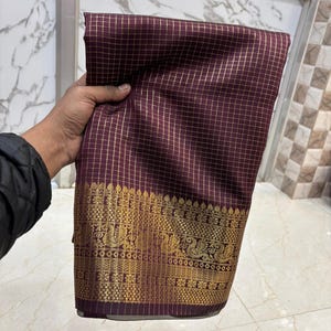 May include: A deep maroon fabric featuring a fine gold grid pattern throughout. The bottom edge is adorned with a wide, intricate gold border displaying repeating peacock motifs and geometric patterns woven into the textile material.