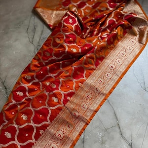 May include: A vibrant orange and red textile with an intricate pattern of floral and geometric shapes. The fabric features gold accents and a detailed border. The textile is draped, showcasing its texture and design.