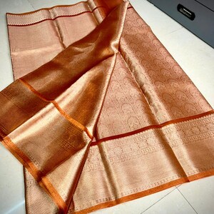 May include: A peach-colored textile with intricate woven patterns and a shimmering, metallic sheen. The fabric features a detailed border with a contrasting orange trim and a darker red accent line. The folds of the fabric create depth and highlight the texture.