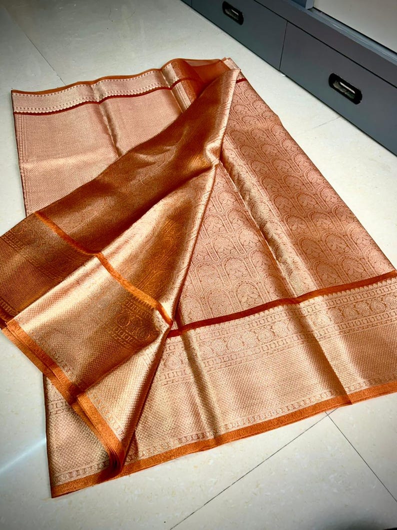 May include: A peach-colored textile with intricate woven patterns and a shimmering, metallic sheen. The fabric features a detailed border with a contrasting orange trim and a darker red accent line. The folds of the fabric create depth and highlight the texture.