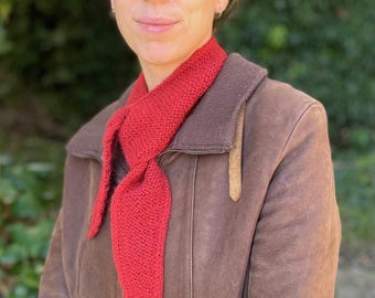 May include: A woman wearing a brown leather jacket with a red knitted scarf tied around her neck. The scarf is a triangular shape with a pointed end.