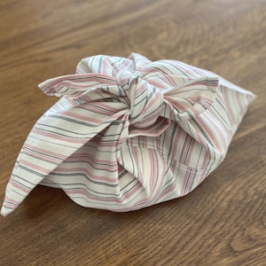 Op de afbeelding: Een witte stoffen tas met roze en zwarte strepen is bovenaan met een knoop vastgemaakt. De tas is gevouwen en lijkt leeg te zijn.