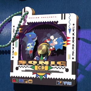 Puede incluir: Un llavero blanco y negro con un disco giratorio que presenta personajes de Sonic the Hedgehog. El llavero tiene una cadena de cuentas verdes y el texto "Sonic the Hedgehog CD" en la parte delantera.