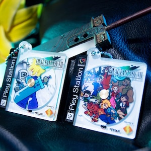 Peut inclure: Deux porte-clés en plastique transparent avec des illustrations de Final Fantasy VII. Le premier porte-clés représente Cloud Strife en bleu et blanc, tenant son épée. Le deuxième porte-clés représente les personnages principaux du jeu en couleur. Les deux porte-clés portent le logo PlayStation et le texte "Final Fantasy VII".