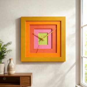 May include: A square wall clock with a layered design in shades of yellow, orange, pink, and green. The clock face features a square-within-a-square design, with gold-colored hands indicating the time. The clock is mounted on a white wall.