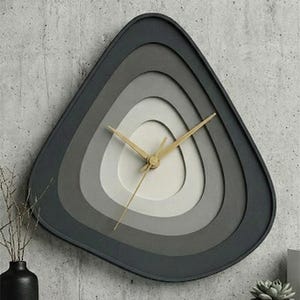 May include: A modern, geometric wall clock with a layered, triangular design. The clock face features concentric rings of gray and white, with gold-colored hands. The clock is mounted on a concrete wall, with a minimalist aesthetic.