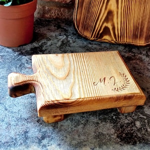 May include: A small wooden serving board with a handle and a decorative design. The board is made of light brown wood and has a dark brown finish. The design is a simple floral pattern with the initials "M.J." in the center. The board is sitting on a gray surface.