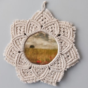 May include: A white macrame mirror with a starburst design and a round mirror in the center. The mirror is hanging from a white string.