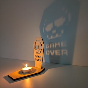 Op de afbeelding: Een houten kistvormige kandelaar met een gepixeld schedelontwerp en de woorden "GAME OVER". Een brandende theelichtkaars werpt een schaduw van een schedel en de woorden "GAME OVER" op de muur. Het thema is gaming.
