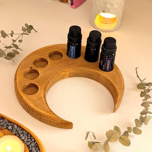 Op de afbeelding: Een halvemaanvormige houten houder voor etherische olie met vijf cirkelvormige inkepingen. Drie kleine zwarte flesjes met etiketten zijn in de houder geplaatst. Op één flesje staat "Eucalyptus" en op een ander "Lavender". De houder is gemaakt van lichtbruin hout.