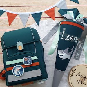 Könnte beinhalten: Ein blaugrüner Rucksack mit Hai- und Ozean-Design. Ein passender blaugrüner Schultüte mit Hai-Design und dem Namen Leon. Die Tüte hat ein weißes Etikett mit der Aufschrift "Schulkind 2024".