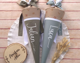 May include: Two cone-shaped school cones with fabric tops featuring alphabet letters. One cone is olive green with the name Julian, the other is grey with the name Luca. Both have white ribbon bows and a wooden disc labelled MariAnna ATELIER.
