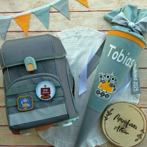 Könnte beinhalten: Ein grauer Rucksack mit Roboter-Patches und ein grauer und orangefarbener Schultüte mit dem Namen Tobias und dem Text "Schulkind 2025".