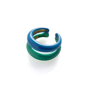 Könnte beinhalten: Zwei gestapelte Ohrklemmen. Die obere Manschette ist glänzend blau und die untere Manschette glänzend grün. Die Manschetten haben ein gebogenes, offenes Design. Der Schmuck ist vor einem weißen Hintergrund platziert.
