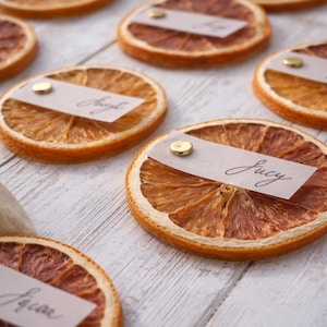 Può includere: Fette di arancia essiccate con biglietti nominativi fissati con spilli dorati. I biglietti sono scritti a mano con nomi come "Lucy" e "Joseph". Le fette sono disposte su una superficie di legno bianco.