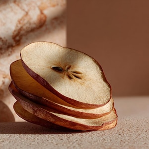 Peut inclure: Gros plan sur des tranches de poires séchées, de couleur brun clair avec des bords brun-rouge. Les tranches sont empilées, révélant la texture du fruit et les détails des graines. Le fond est de couleur neutre et douce.
