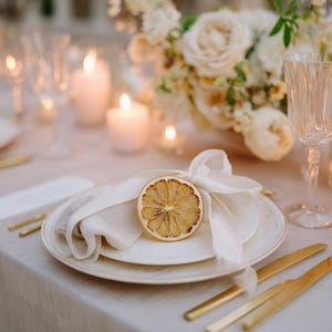Peut inclure: Mise en place élégante avec des assiettes blanches, une serviette blanche pliée et une tranche de citron séché attachée avec un ruban blanc. Couverts dorés et composition florale avec bougies.