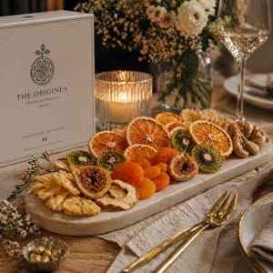 Peut inclure: Une boîte rectangulaire étiquetée THE ORIGINEA ARTISANAL PRODUCTS FRANCE COLLECTION DE FRUITS SECHES est posée à côté d'un plateau en marbre garni de tranches d'orange séchées, de kiwis séchés, de figues et d'abricots secs sur une table.