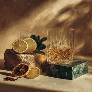 Peut inclure: Un verre à whisky en cristal rempli de glace et de liquide ambré, posé sur un bloc de marbre vert. Des tranches de citron, des tranches d'orange séchées et des feuilles de menthe sont disposées sur une pierre.
