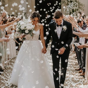 Peut inclure: Un couple de mariage marchant ensemble sous une pluie de pétales de fleurs blanches. La personne à gauche porte une robe à corsage en dentelle blanche avec une jupe en tulle et tient un bouquet de roses blanches. L'autre porte un costume noir.