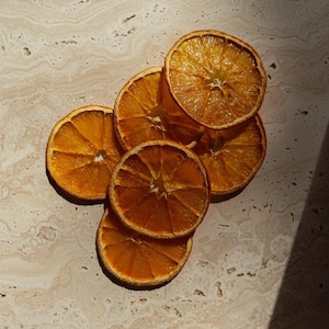 Könnte beinhalten: Nahaufnahme von mehreren getrockneten Orangenscheiben, die auf einer hellen, strukturierten Oberfläche angeordnet sind. Die Scheiben haben eine warme, goldorange Farbe mit sichtbaren Texturen und Details. Das Bild ist gut beleuchtet.