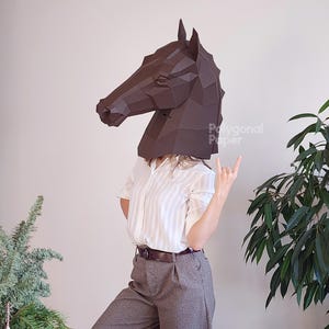 May include: A dark brown, geometric horse head mask. The mask is made of paper and covers the head. The person is wearing a white striped shirt and gray pants. The person is giving the rock on hand gesture.