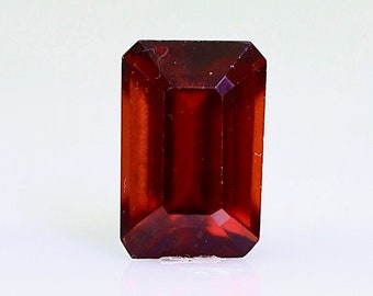 May include: A deep red gemstone with a rectangular emerald cut. The gemstone is faceted and has a high polish.