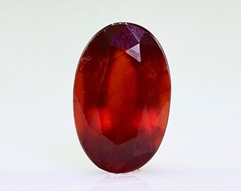 May include: A faceted, oval-shaped gemstone with a deep red hue. The gem is set against a white background, showcasing its polished surfaces and internal brilliance. The gemstone's facets catch the light, creating a play of color and sparkle.