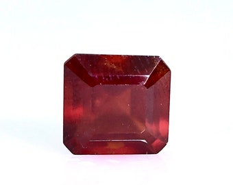 May include: A deep red gemstone with a square emerald cut. The gemstone is faceted and has a slight shine.
