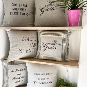 May include: Four decorative throw pillows with inspirational Italian phrases printed on them. The pillows are beige with black text and are displayed on a wooden shelf. The phrases are "Se puoi sognarlio puoi farlo", "Oggi scelgo la Gioia", "Dolce Far Niente", and "Oggi scelgo la Gioia".