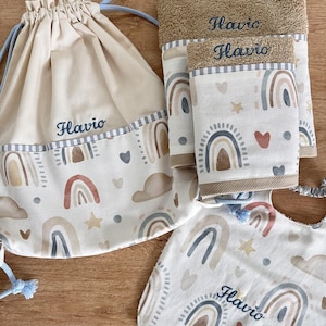 Op de afbeelding: Een set beige babyartikelen met een regenboog- en wolkenpatroon. De set bevat een trekkoordtas, handdoeken en een slab, allemaal geborduurd met de naam "Flavio". De handdoeken zijn lichtbruin.