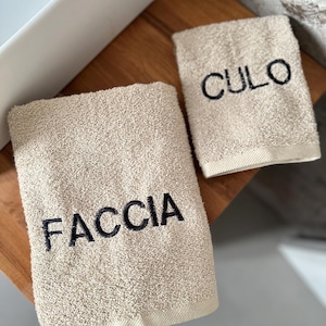May include: Two beige bath towels with black embroidered text. One towel reads "FACCIA" and the other reads "CULO". The towels are made of a soft, absorbent material and are likely used in a bathroom setting.