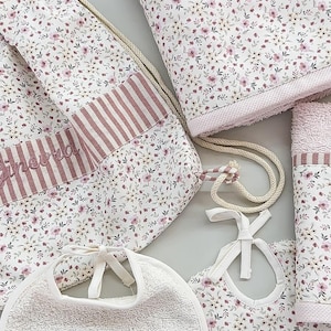 Könnte beinhalten: Ein Baby-Geschenkset mit Lätzchen, Handtuch und Kordelzugbeutel. Die Artikel sind weiß mit floralem Muster und rosa Akzenten. Der Name "Ginevra" ist rosa auf Lätzchen und Handtuch gestickt.