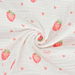 Könnte beinhalten: Ein weißer Musselinstoff mit einem sich wiederholenden Muster aus roten Erdbeeren mit grünen Blättern und kleinen roten Herzen. Das leichte Textil ist in einer zentralen Spirale gerafft und zeigt die weiche, geknitterte Struktur des Materials.