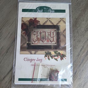 May include: A Just Nan Ginger Joy 2006 Christmas Ornament cross stitch pattern kit in a clear plastic bag. The cover features a stitched design with the word JOY, a small tree, and a figure. A small bag of beads and a charm are included inside.