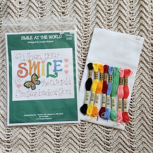 May include: Cross-stitch kit with the text "When you SMILE the World Smiles back at You." The kit includes a printed pattern, embroidery floss in various colors, and a piece of white Aida cloth. The design features a butterfly and hearts.