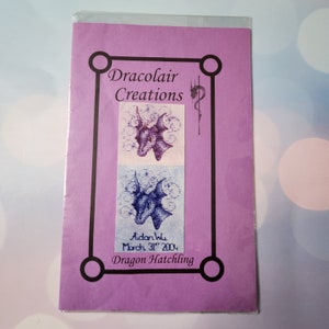 May include: A lavender-colored package featuring a cross-stitch pattern of a dragon's head. The package has the text "Dracolair Creations" and "Dragon Hatchling". The dragon is depicted in purple and blue.