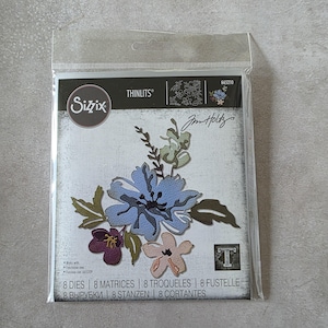 May include: A package of Sizzix Thinlits dies featuring floral designs. The dies include a large blue flower, a small pink flower, and other foliage. The package also includes the text "Jim Holtz" and "8 DIES | 8 MATRICES | 8 TROQUELES | 8 FUSTELLE | 8 ВЫРУБКИ | 8 STANZEN | 8 CORTANTES".