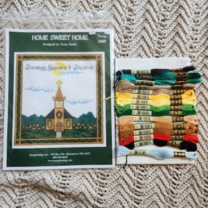 May include: A cross-stitch kit featuring a church design with the words "Home Sweet Home." The kit includes a printed pattern, embroidery floss in various colors, and a piece of Aida cloth. The kit is designed by Tracy Fuchs.