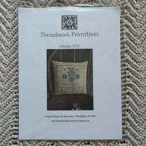 May include: A white paper with the text "Threadwork Primitives" and "Winter 1737". The image on the paper shows a cross-stitch design of a pillow with winter-themed motifs and the word "Winter". The bottom of the paper has the text "Original Designs by Nan Lewis - Philadelphia, PA 19111".