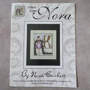 May include: A cross-stitch pattern titled "Letters From Nora" by Nora Corbett. The design features a fairy with a letter "K" in a decorative frame. The background is white with a rose pattern.