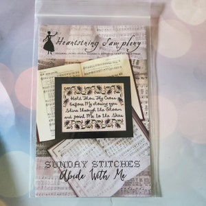 May include: A cross-stitch pattern packet titled "Abide With Me" by Heartstring Samplery. The design features a framed cross-stitch with the text "Hold Thou Thy Cross" and floral accents. The background includes sheet music.