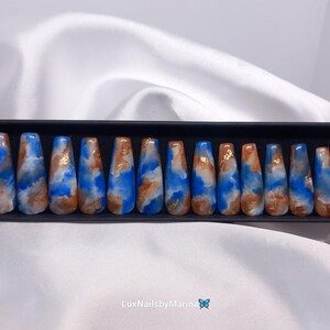 Può includere: Un set di 20 unghie finte press-on blu, bianche e dorate con un design a spirale in una scatola nera. Le unghie sono lunghe e hanno un design marmoreo.