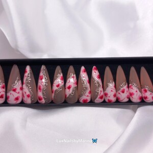 Può includere: Un set di 20 unghie finte in una scatola nera. Le unghie sono lunghe e a forma di stiletto. Sono dipinte con una base marrone chiaro e un design floreale bianco e rosso. Le unghie sono disposte in due file da dieci.