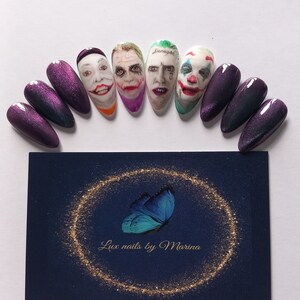 Pode incluir: Conjunto de dez unhas postiças com acabamento de glitter roxo. Cinco das unhas apresentam imagens pintadas à mão do Joker do filme "Joker". As unhas estão dispostas em semicírculo sobre uma superfície branca. Um cartão de visita com o texto "Lux nails by Marina" está localizado no centro inferior da imagem.