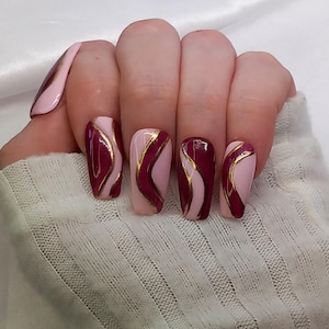 Può includere: Primo piano di una mano con unghie lunghe di colore bordeaux e rosa con accenti dorati. Le unghie sono decorate con un design ondulato.