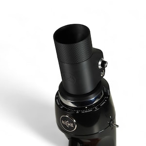 May include: Black coffee grinder with a textured top and a dial for grind size. The grinder has the word "NiCHE" on the front and is set against a white background. The grinder is designed for filter coffee.